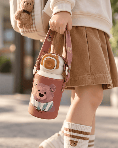 A child holds a pink water bottle with a cute teddy bear design and a matching shoulder strap. The child is wearing a cream sweatshirt, a brown corduroy skirt, and striped socks, with a matching backpack visible in the background.