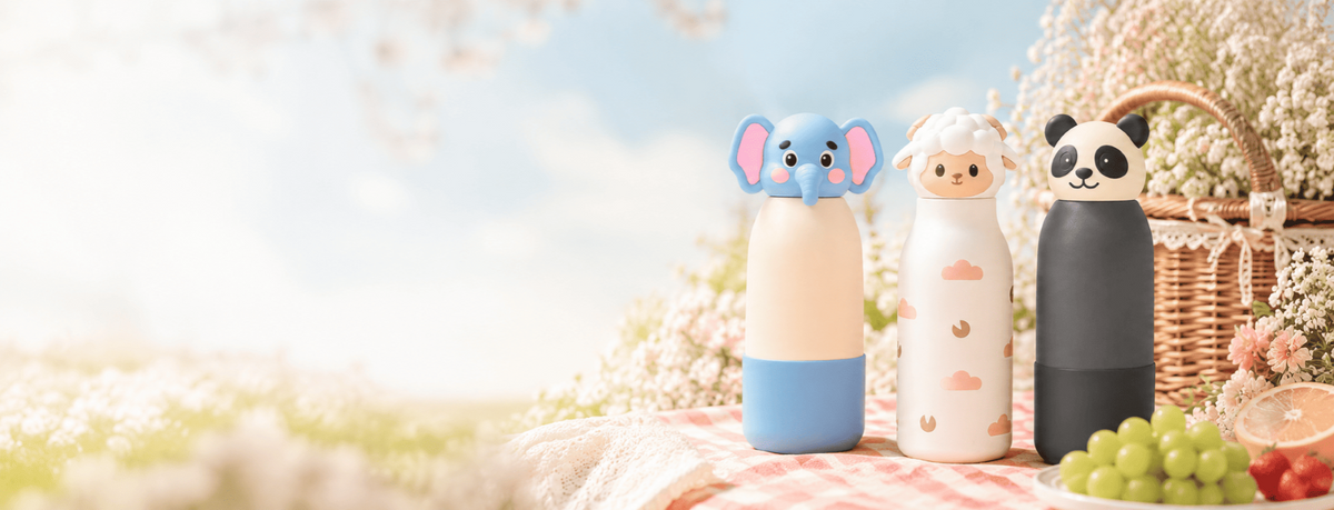 Three cute animal-shaped insulated bottles with matching lids are lined up on a picnic blanket outdoors. The bottles feature elephant, sheep, and panda designs in pastel colors.